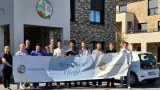 Gruppenfoto vor einem modernen Seniorenwerk-Gebäude: zwölf Mitarbeitende und Auszubildende halten ein großes Banner mit der Aufschrift „Wir leben Pflege.“; rechts steht ein weißes Dienstfahrzeug, sonniger Tag.