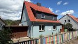 Das zum Verkauf stehende Gebäude in Herrmannsacker: ein zweigeschossiges Wohnhaus mit hellblauer Fassade und rotem Satteldach. Davor ein bunt gestrichener Lattenzaun, links ein Carport. Im Hintergrund Bäume und blauer Himmel.