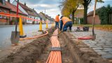 Bauarbeiten in einer Dorfstraße in Herrmannsacker: orangefarbene Glasfaserkabel liegen in einem offenen Graben, Absperrbaken sichern die Baustelle. Die Szene zeigt den Breitbandausbau der Deutschen Telekom und symbolisiert den Fortschritt der digitalen Infrastruktur in Harztor. KI