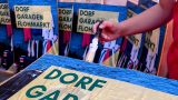Eine Person streicht ein Plakat mit der Aufschrift „Dorf Garagen Flohmarkt“ ein; im Hintergrund sind mehrere identische Plakate zu sehen.