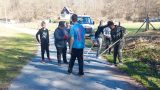 Gruppe Jugendlicher bessert bei sonnigem Wetter einen Weg mit Schotter aus; einige arbeiten mit Rechen, andere stehen zusammen und unterhalten sich. Im Hintergrund ein Transporter und ein bewaldeter Hang.