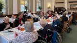 Bewohnerinnen und Bewohner eines Pflegeheims sitzen in einem festlich gedeckten Speisesaal an langen Tischen und genießen ein gemeinsames Herbstfest. Einige Gäste sitzen in Rollstühlen, andere unterhalten sich oder klatschen Beifall. Auf den Tischen stehen kleine Kürbisse und herbstliche Dekorationen, im Hintergrund spielt ein Musiker. Die Atmosphäre wirkt warm und gesellig.