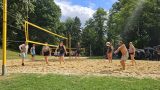 Beachvolleyball auf dem Sandfeld: gemischtes Team im Spiel, Spieler rechts baggert den Ball; Sonnenschein, Bäume und Zuschauerzelte im Hintergrund.