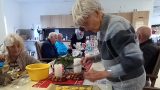 Mehrere Senioren sitzen in der Tagespflege an einem Tisch und bereiten Plätzchen zu. Eine Frau bedient einen Fleischwolf, eine andere verziert die ausgestochenen Plätzchen auf dem Blech. Auf dem Tisch stehen Kerzen und weihnachtliche Dekoration.
