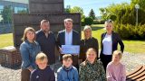Eine Gruppe von Erwachsenen und Kindern steht bei sonnigem Wetter vor einem Denkmal im Freien. Ein Mann in der Mitte hält einen symbolischen Spendenscheck in den Händen. Alle lächeln in die Kamera.
