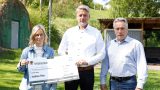 Drei Erwachsene stehen im Freien und halten gemeinsam einen symbolischen Spendenscheck in die Kamera; im Hintergrund sind Bäume, Rasen und ein kleines Gebäude zu sehen.