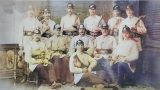Historisches, handkoloriertes Gruppenfoto: rund ein Dutzend uniformierte Feuerwehrmänner in hellen Jacken und Helmen, mit Lederkoppeln, Äxten und Werkzeugen; Studioaufnahme – einige sitzen, andere stehen, einer liegt vorn auf einem Teppich (um 1900).