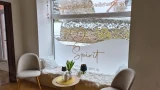 Ein Fensterplatz mit zwei grauen Sesseln und einem kleinen Tisch. Die untere Hälfte der Fensterscheibe ist milchig foliert und trägt den goldenen Schriftzug 'Spirit - Zentrum für ganzheitliche Heilbehandlungen'. Auf der Fensterbank liegen flauschige weiße Kissen.