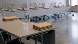 Blick in einen modernen Sitzungssaal vor einer Sitzung. Helle Konferenztische stehen in U-Form auf grauem Fliesenboden. Darauf liegen gestapelte Aktenmappen, dazu Wasserflaschen und Gläser. Im Hintergrund leere graue Stühle und hellgraue Wandpaneele. KI.
