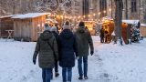 Verschneiter Park an einer alten Klosterkirche am Abend. Kleine Holzstände sind mit Lichterketten geschmückt. Besucher in Winterjacken gehen über den Markt. KI.