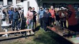 Eine Gruppe überwiegend älterer Menschen steht bei Sonnenschein auf einer Wiese, einige halten Nordic-Walking-Stöcke. Rechts legt ein Mann mit roter Kappe und Weste ein Lasergewehr an. Im Vordergrund stehen Holzbänke und ein Tisch.