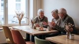 Gemütlicher Café-Raum mit Holztischen und Trockenblumen-Deko, in dem mehrere ältere Menschen bei Kaffee und Kuchen miteinander ins Gespräch vertieft sind. KI.
