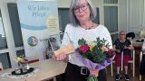 Renate Völker steht in einem hellen Aufenthaltsraum des Seniorenpflegeheims Sonnenhof. Sie hat graue, schulterlange Haare und trägt eine schwarze Brille, eine weiße Bluse mit schwarzen Punkten und einen schwarzen Rock. In der rechten Hand hält sie einen bunten Blumenstrauß, in der linken eine gläserne Auszeichnung auf einem Holzsockel. Hinter ihr steht ein Roll-up mit der Aufschrift „Wir leben Pflege." und dem Logo des Seniorenwerks. Im Hintergrund sitzen zwei ältere Bewohnerinnen an Tischen.