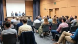 Das Bild zeigt eine große Gruppe von Menschen, die in einem Konferenzraum sitzen und einer Reihe von Personen zuhören, die auf einer Bühne stehen. Die Personen auf der Bühne scheinen eine Präsentation oder eine Rede zu halten. Der Raum ist gut beleuchtet und die Atmosphäre scheint formell zu sein.