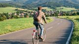 Eine Person faehrt mit einem gewoehnlichen Fahrrad in Alltagskleidung auf einer ruhigen Landstrasse im Harz. Es ist Tag, das Wetter ist freundlich. Das Gesicht ist nicht erkennbar. KI.