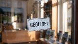 Geöffnet-Wendeschild an der Glastür: Ein kleines Wendeschild hängt an einer Kette hinter einer Glastür. Auf der sichtbaren Seite steht „Geöffnet" in schwarzer Schrift auf weißem Grund. Durch die Glastür fällt Tageslicht. Im Hintergrund ist verschwommen ein Innenraum zu sehen. KI.