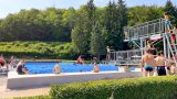 Menschen im Freibad vor bewaldeter Kulisse – Besucherinnen und Besucher schwimmen, sitzen am Beckenrand oder stehen am Sprungturm. Sommerliche Szene im Waldbad Ilfeld der Gemeinde Harztor. Gesichter wurden unkenntlich gemacht.